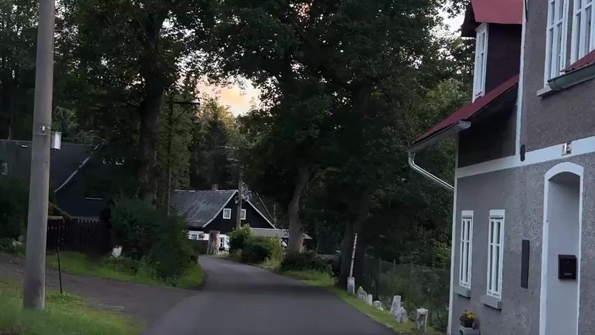 A rural road winds through a village surrounded by trees. On the right side of the road is a grey house with white trim and a red roof. On the left side, several smaller houses are visible, also surrounded by trees. The sky in the distance is a soft orange, suggesting either sunrise or sunset. A power pole stands on the far left of the frame. The scene is quiet and appears to be undisturbed.