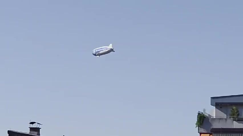 The video shows an aerial view of a white blimp with blue stripes flying in a clear blue sky. Below the blimp, the tops of modern houses with peaked roofs and satellite dishes are visible. Green trees and bushes surround the houses, and a grassy field is in the foreground. The blimp appears to be moving from left to right across the frame.