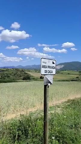 The video shows a landscape with a field of tall, green grass under a bright blue sky dotted with white clouds. In the foreground, there is a wooden post with a sign that reads "AREA PRIVADA DE CAÇA" (Private Hunting Area) in white letters on a black background. Beyond the field, there are rolling hills with some trees and a distant view of buildings and an industrial area. An electrical transmission tower is visible in the distance. The camera pans slightly, revealing more of the field and the surrounding vegetation.