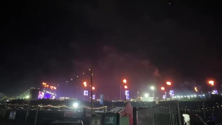 A nighttime long shot shows a wide outdoor music festival stage with many glowing lights. The stage, visible on the left, displays vibrant blue and purple lights. Strings of lights are visible, with some lights suspended vertically from poles on either side of the shot. A smoky cloud is visible in the middle of the frame, with other smaller puffs of smoke visible to the right. Beyond the lights, several tents are visible in the background.