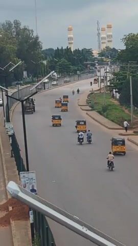 A slightly elevated, medium shot captures a street scene in what appears to be a city. The road is paved and curves gently to the left. Several yellow, three-wheeled auto-rickshaws, commonly known as Keke Napeps, are visible on the road, as well as two motorcycles with riders, and a few cars. On the right side of the road, a paved sidewalk runs parallel, with pedestrians walking. Beyond the sidewalk, there is lush green vegetation and trees. In the background, tall, slender white structures with golden domes rise above the trees, resembling minarets of a mosque. The sky is overcast with a cloudy, pale gray hue. The foreground features a section of a white metal railing, possibly from a bridge or overpass, and a solar-powered street light. A poster is attached to a pole on the left side of the road. The overall impression is of a busy urban environment with a prominent religious structure in the distance.