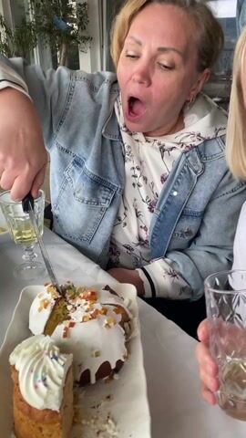 Two middle-aged, blonde women are seated at a table covered with a white tablecloth. A platter of cake sits in front of them. The woman on the left, wearing a denim jacket and a floral hoodie, is cutting the cake with a knife. She has her mouth open in a wide yawn or mock eating pose. The woman on the right, dressed in a white sweater with a Union Jack design, is holding a glass of a clear liquid and looking at the other woman. In the background, through the windows, hints of a landscape can be seen. The atmosphere is casual and possibly celebratory.