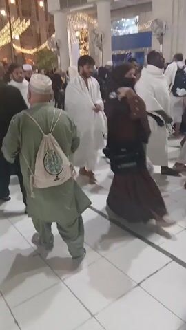 The video shows a crowd of people walking in a plaza. Many of the men are wearing white robes, suggesting they may be pilgrims.  Some people are wearing more casual clothing. There is a cleaning machine parked on the side. The plaza is lined with buildings, some of which are decorated with lights. The overall atmosphere appears to be busy and communal.