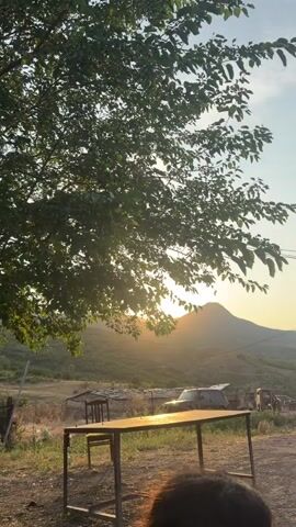The video opens with a shot of a leafy tree obscuring the sun, which is low in the sky and casting a warm glow. The sun is behind a mountain in the distance. In the foreground, there is a wooden table and chair. A car with its trunk open is parked behind the table. A person walks from right to left across the frame in the mid-ground. The scene appears to be outdoors in a rural or semi-rural setting, possibly during sunset or sunrise.