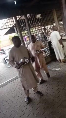 The video shows several men walking through what appears to be an outdoor market or gathering place. They are dressed in various styles of clothing, including traditional robes. Some of the men are carrying plates or containers of food. The environment is slightly cluttered, with wooden structures and other objects visible in the background. The camera appears to be moving along with one of the individuals.
