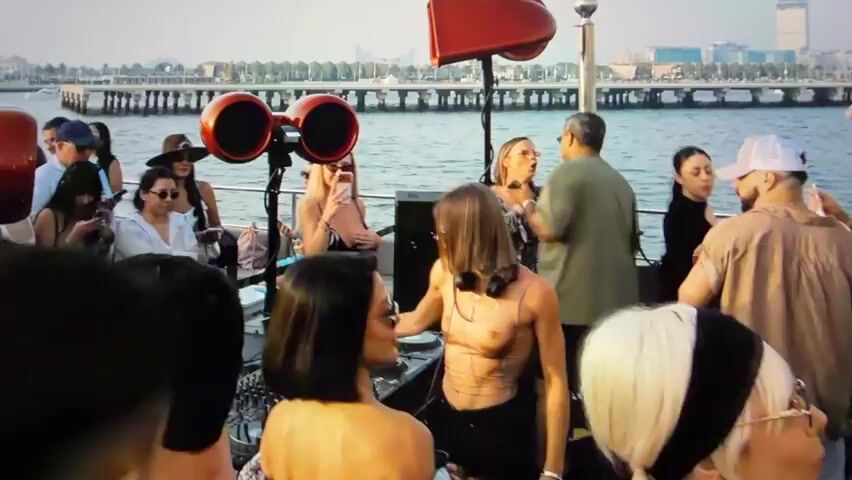 A group of people are on a boat party. A DJ is seen at the front of the boat with music equipment. People are dancing and socializing. In the background, there is a body of water with a cityscape and a pier.