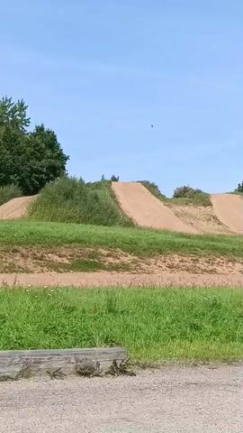 The video pans across a dirt track with several jumps and berms, surrounded by grass and trees under a clear blue sky. There are no people or vehicles visible in the footage. The track appears to be an outdoor motocross or BMX course.