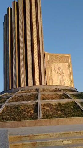A low-angle shot captures a monument with a tall, layered structure resembling stacked pillars. The pillars are light brown with darker brown stripes and cast shadows. To the right of the pillars is a rectangular panel with a bas-relief sculpture of a person in profile, wearing a robe and with their hands raised. The panel is framed by an ornate, gold-colored border with Arabic script. The monument sits on a circular, tiered base covered in dry grass and bordered by a metal grid. In the foreground, stone steps lead up towards the base. The sky is a clear, pale blue. The sun illuminates the scene from the right, creating highlights and shadows.