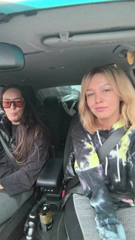 Two women are in the front seats of a car, wearing seatbelts. The woman on the left has long dark hair and is wearing sunglasses and a black puffy vest. The woman on the right has short blond hair and is wearing a tie-dye hoodie. They are both facing forward and appear to be talking. There is a drink in the center console. It's daytime, and the background appears to be a residential area.