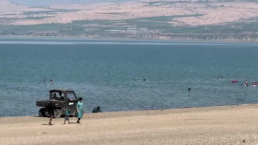 A wide shot shows a beach with people walking and swimming in the sea. In the foreground, three people are walking on the beach, and a small, black vehicle is parked on the sand near the water's edge. In the background, the sea stretches out to meet a distant shore with hills and vegetation. Several people are visible swimming and playing in the water. The sky is clear and the weather appears to be sunny.