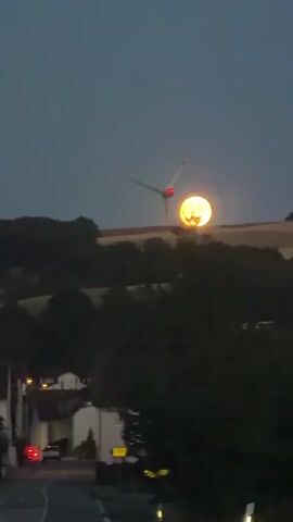 The video captures a view of a rural landscape at dusk or dawn. A wind turbine stands prominently on a hilltop, silhouetted against a large, bright, and possibly setting or rising sun or moon. The sun/moon is so bright it appears to have details within it. Below the hill, a small village is visible with some lights illuminated. A road leads into the village, and trees obscure parts of the scene. The sky is a muted blue, indicating twilight. There are no people or animals clearly visible, and no apparent actions or interactions are taking place.