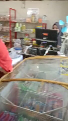The video depicts an indoor scene, possibly a small convenience store or market. In the foreground, a person wearing a red top and dark pants is visible from the waist down. To the left of this person, a person wearing a black head covering and a grey dress is standing. In the background, a young child wearing a red and yellow outfit is interacting with another person. Shelving units filled with various products are visible behind the child and the other person. A glass display case or window is present on the right side of the frame, showing more items inside. The flooring appears to be tiled. The overall lighting suggests an interior setting with artificial light sources.
