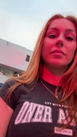 The video is a selfie taken by a young woman with red hair who is looking directly at the camera. She is wearing a dark t-shirt with white text that says "ONER AD." She is illuminated by red light. In the background, there is a crowd of people watching a large screen that is displaying a soccer match. Some people are wearing jerseys and scarves in team colors. The environment appears to be an outdoor event or viewing party. The woman shifts her gaze slightly away from the camera in the middle of the clip. The background blurs in and out of focus.