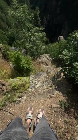The video begins with a first-person perspective looking down at the feet of a person wearing sneakers and blue jeans, standing on a dirt and rock surface with small plants. The view immediately below shows a steep drop-off into a forested valley. The camera then slowly pans upward, revealing a vast, deep, and heavily forested mountainous valley or gorge under a blue sky with scattered white clouds. Sunlight is visible, creating a bright illumination over the natural landscape.
