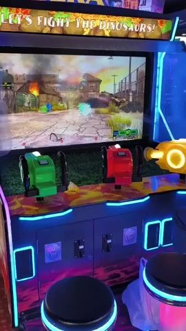 The video shows an arcade game called Dinosaur Era, where players use toy guns to shoot at dinosaurs on a screen. The game has a futuristic design with neon lights and a vibrant color scheme. The screen displays a scene of a city under attack by dinosaurs, with explosions and fire. The players are not visible, but the game controllers, two green and one red, are positioned in front of the screen, indicating that at least two people are playing. A yellow toy gun is also attached to the side of the machine. The environment appears to be an arcade, with other game machines visible in the background. The overall impression is one of excitement and action, characteristic of arcade games.