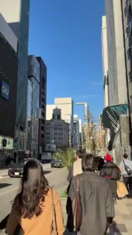 The video shows a sidewalk scene in a city. Several people are walking along the sidewalk, with traffic passing on the street. Buildings line both sides of the street, including one with the "GU" logo visible. A building with a distinctive tower is visible in the distance. The sky is clear and blue.