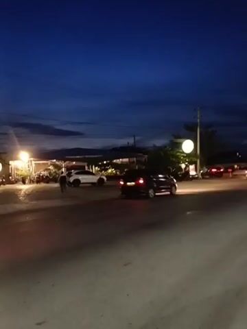 The video opens at dusk. A road is in the foreground, and to the left, there is a restaurant with bright lights. Several cars are parked outside the restaurant and on the road. A person is seen walking away from the camera on the left side of the road. A white SUV is parked to the left of the restaurant. In the background, there are some buildings and trees. The sky is a deep blue, with some clouds. The video shows a red car driving past in the foreground.