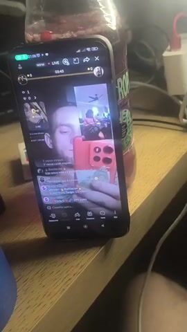 The video shows a close-up view of a smartphone screen displaying a live video call. The main window on the phone screen features a person's face looking directly at the camera, holding another phone that reflects a pink phone case. A smaller window on the top right of the screen shows another person in what appears to be a room with a ceiling fan. Below the main video window, there are various icons and text elements typical of a social media or video calling application, including viewer counts, comments, and interaction buttons. The phone is placed on a wooden surface, possibly a desk, next to a dark object on the left, and a bottle with a red liquid and green label on the right. A keyboard is partially visible in the background. The lighting suggests an indoor environment, possibly at night.