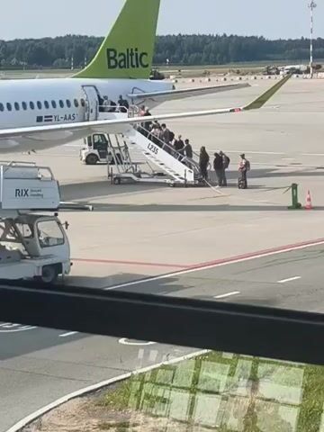 The video shows an airplane parked on an airport tarmac. Passengers are boarding the plane using a mobile staircase. The plane has "Baltic" written on its tail fin and the registration number YL-AAS is visible on the fuselage. Ground handling equipment, including a baggage loader and a pushback tractor, are parked near the plane. The scene appears to be a sunny day with a clear sky. The video is filmed from an elevated perspective, likely from inside another aircraft or an airport terminal.