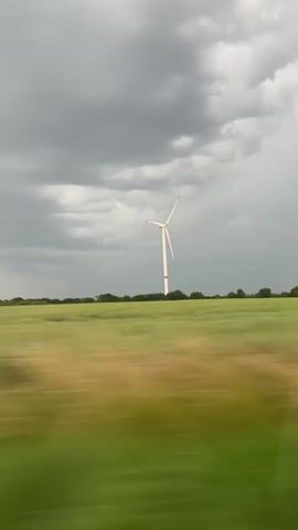 A video shows a wind turbine in a field under a cloudy sky. The camera appears to be moving, causing a blur in the foreground of the green field. The wind turbine is white with a red band near the top of its tower and has three blades.