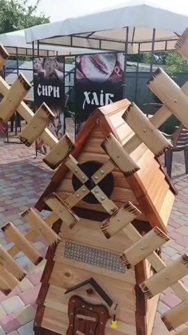 The video shows a close-up of a wooden windmill decoration placed on a patterned paved surface. The windmill is detailed with a triangular roof, several blades made of wooden planks attached to a central cross-shaped structure, and a small door-like feature at the base. Behind the windmill, a partially visible gazebo structure with a white canopy is present, along with a fence, tables, and chairs. Posters or signs with text, possibly in Cyrillic script, and images of bread and other food items are visible on the fence. The lighting suggests it is daytime, with natural light illuminating the scene. The focus is on the windmill, which appears to be stationary.