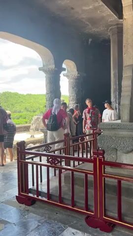 The video opens with a wide shot of an outdoor historical site, likely a tomb or temple. In the foreground, a vibrant red and gold barrier separates the viewer from a stone monument. Behind the barrier, several tourists are gathered, looking at the monument and the surrounding architecture. The monument itself is large and intricately carved, with a tall stone slab. The architecture features large, weathered stone pillars and arched doorways. Lush green trees and foliage are visible in the background, suggesting a natural, possibly tropical environment. The lighting indicates it is daytime, with sunlight filtering through the structures. One person in the foreground wears a pink headscarf and a backpack, while another wears a brightly colored floral shirt. The other individuals appear to be tourists, some looking around, others interacting with each other. The camera pans slightly, revealing more of the monument and the surrounding area, hinting at the scale and grandeur of the historical site.