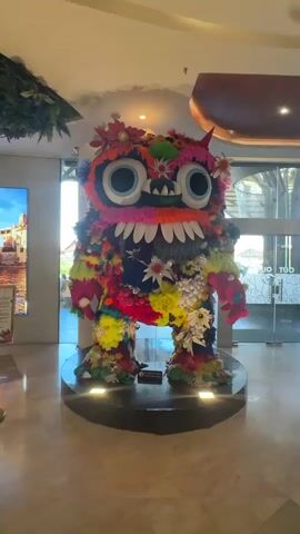 A colorful, fuzzy, monster-like statue with large blue eyes and a wide mouth with white teeth stands on a black circular platform. The statue is adorned with various colorful flowers, pom-poms, and yarn in shades of pink, blue, yellow, orange, green, and white. The monster has two small red horns on its head and furry arms and legs. The background appears to be an indoor public space, possibly a mall or lobby, with a marble floor, a large digital display on the left, and glass doors in the distance. Lighting from below illuminates the base of the statue.