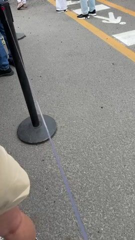 The video shows a group of people standing behind a black post with a rope barrier. The group appears to be waiting in line. They are standing on a paved surface marked with yellow lines. In the background, there is a rocky hill with sparse vegetation. Some people are standing on top of the hill. The sky is cloudy. Several cars are parked nearby.
