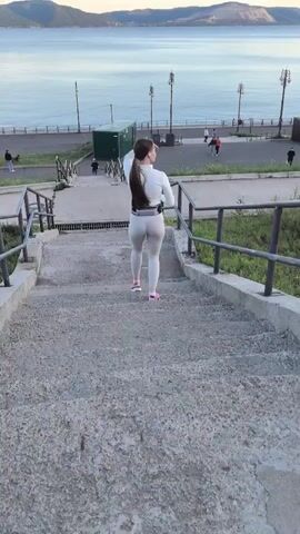 The video shows a woman with a ponytail walking up a concrete staircase. She is wearing a white athletic top and light-colored leggings, with a small white pouch around her waist. Her sneakers are pink. The camera is positioned behind her, looking up the stairs.

The staircase is outdoors, with metal railings on either side. Green grass and some bushes are visible along the sides of the stairs. In the background, there is a body of water, likely a river or a large lake, with hills or mountains on the opposite shore. The water is calm and reflects the sky. Along the waterfront, there is a paved walkway with lampposts and benches. Several people are visible walking or standing on the walkway in the distance. A small green building or structure is also present near the top of the stairs.

The scene appears to be set during either sunrise or sunset, as the lighting suggests the golden hour. The sky is a soft orange and blue, indicating pleasant weather. The overall atmosphere is peaceful and suggests an individual engaged in a physical activity like jogging or walking for exercise, enjoying the scenic view. No text is visible in the media file.