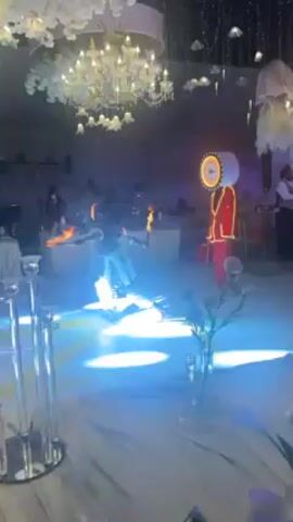 The video shows a fire performer and a person dressed in a drum costume performing at an event. The fire performer, dressed in a black and white costume, spins flaming batons.  A person in a red drum costume stands nearby with lights outlining the drum.  They are on a light tan dance floor. There are tables with decorations and people in the background and a chandelier hanging from the ceiling. Lighting effects alternate between white and blue.