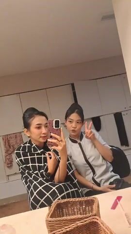 Two young women are seen in a mirror selfie video, likely taken in an indoor setting such as a dressing room or a casual room. The woman on the left, wearing a black and white checkered garment and with her hair pulled back, holds a smartphone with a distinctive patterned case, capturing their reflection. She makes various playful expressions, including sticking out her tongue, putting a finger to her lips, and smiling. The woman on the right, wearing a light-colored uniform-like top and with her hair also pulled back, joins in with playful poses like a peace sign and sticking out her tongue.

In the foreground, on a table, there are two wicker baskets, a red lipstick or marker, and a partially visible box with the word "JUMBO" on it. The background features white cupboards or lockers, with some clothing or towels hanging, and a black chair. The overall mood is lighthearted and playful. The lighting is artificial, indicating an indoor environment. Spoken words, possibly in Indonesian, can be heard in the background, including "Đã là bà" and "Bye." The location context is Singkawang, Indonesia.