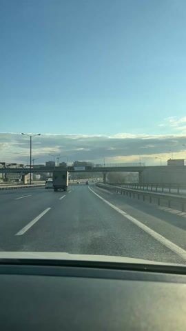 The video shows a view from inside a moving vehicle driving on a highway, the road surface is wet. The sky is clear and blue with some thin cloud coverage.  A grey truck is in the adjacent lane to the left. Above the highway is an overpass with a large billboard advertising Dice Kayek timepieces. In the distance, there are buildings and streetlights. The car is following the lanes and maintaining a steady speed.