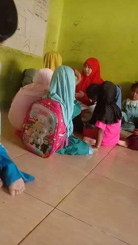 A group of children are sitting on the floor in what appears to be a classroom or common area. The walls are a bright green, and the floor is tiled in a neutral tone. Several children are wearing hijabs in various colors, including red, yellow, and turquoise. One child in the foreground is wearing a pink hijab and has a pink backpack adorned with cartoon characters. Another child, wearing a pink shirt, is kneeling. In the background, a child in a light blue outfit is holding a phone. The children seem to be interacting with each other or observing something. The overall impression is one of a casual gathering or learning activity.