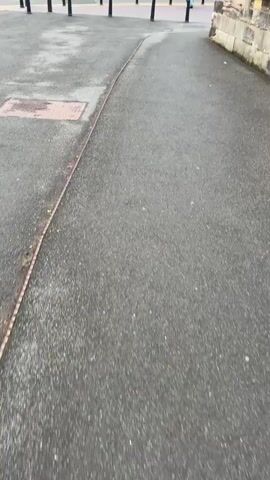 The video shows a person walking in what appears to be a public square or street in Ironbridge, United Kingdom. The scene is dominated by a large, old brick building with numerous windows, some of which are adorned with green shutters. A church steeple is visible in the background, situated on a hill above the brick building. The ground is paved with asphalt, and several black bollards are arranged in a line across the area.

The person in the video is wearing light-colored clothing. They are walking away from the camera and towards the brick building. The weather appears overcast, suggesting it might be a cloudy day. The lighting indicates it is likely daytime, possibly late afternoon or early evening. There is a sign above the entrance of the brick building with some text on it, but it is not legible. The overall atmosphere is calm and ordinary, depicting a person going about their day in this historic location.