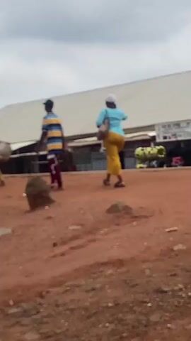 In Jere South, Nigeria, two individuals are captured in a brief visual moment. A young person, adorned in a horizontally striped t-shirt and dark pants, walks away from the camera, pulling a large, woven basket. To their right, a woman walks in the same direction, wearing a blue t-shirt and a long, flowing yellow skirt. She carries a light-colored bag over her shoulder.

The scene unfolds on a dusty, reddish-brown path, indicative of an outdoor, likely rural or semi-urban environment. The ground is uneven, with scattered rocks and debris. In the background, a long, white-roofed building stretches across the frame, suggesting a market or a series of shops. Some stalls are visible beneath the roof, with what appear to be goods displayed. The sky above is overcast, hinting at cloudy weather.

The activity depicted is simple: two people walking, possibly on their way to or from a marketplace or performing daily errands. There are no overt displays of emotion, but their posture suggests a steady, unhurried pace. The time of day is not precisely discernible due to the cloudy conditions, but the light seems muted.

Visible text includes a sign on the background building with lettering in a language likely indigenous to Nigeria, though it is not clearly legible. The overall impression is one of everyday life in a Nigerian community, with a focus on the movement of people and their engagement with their surroundings.