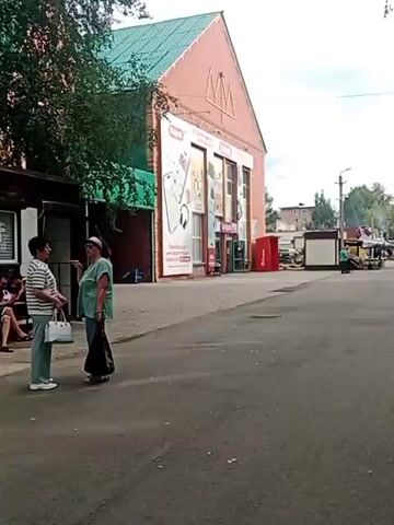 Two people are standing on a sidewalk in front of a building with advertisements on it. One person is wearing a striped shirt and the other is wearing a green shirt. There are trees in the background.