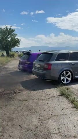 The video shows a gray Audi station wagon parked on a dirt road next to a purple Volkswagen Golf. Both cars are modified with aftermarket wheels and rear spoilers. There is exhaust smoke coming from the exhaust of the Golf. The background includes a green tree, other cars, and a blue sky with scattered clouds. Mountains can be seen in the distance. The environment appears to be an outdoor event with vehicles present.