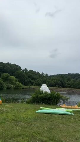 The video pans across a grassy area with several kayaks lying on the ground. In the background, a lake is visible, with a wooded area beyond it. A large white inflatable object is situated near the lake. Further to the right, people are seen interacting with inflatable water structures in the lake. Some individuals are swimming or playing on the inflatables, while others are standing on the shore. A wooden building and a row of benches are also visible. The sky is overcast.