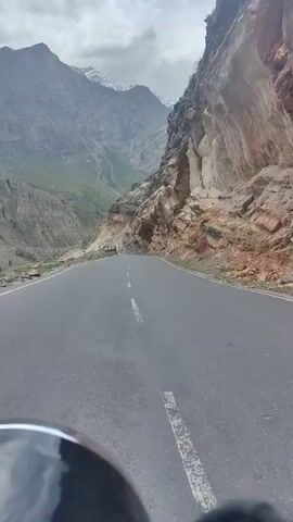 The video shows a first-person perspective of someone riding a motorcycle on a winding mountain road. The road is paved and has white dashed lines dividing the lanes. On the right side of the road, a steep, rocky cliff face dominates the landscape. The cliff shows signs of erosion and loose rocks. On the left side, the road drops off into a deep valley with a river or stream visible at the bottom. Further up the valley, more mountains rise, some with snow-capped peaks, under a cloudy sky. The motorcycle's handlebars and part of the seat are visible in the foreground, indicating the rider's viewpoint. The rider is in motion, traveling forward along the road. There are no other vehicles or people clearly visible on the road itself, although distant figures or structures might be present in the valley. The overall environment is mountainous and appears to be a remote or scenic route.