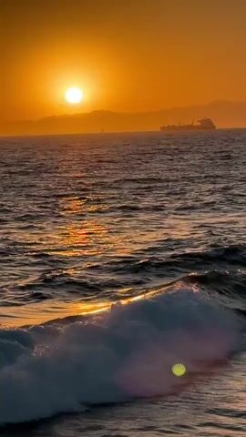 A video captures a sunset over a body of water. The sun is a bright, round orb low in the sky, casting a warm orange glow across the scene. In the distance, mountains form a silhouetted horizon. A large ship is visible on the water, its shape dark against the vibrant sky. The water itself is choppy, with waves cresting and breaking, reflecting the sunlight in shimmering patterns. The foreground features a breaking wave, its white foam contrasting with the darker water. The overall atmosphere is serene and picturesque.