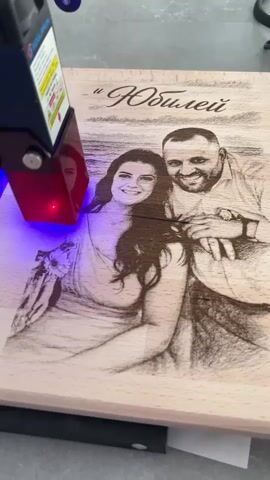 A laser engraver is in operation, focused on a wooden surface. The laser beam emits a red light. The wooden surface is engraved with a portrait of a couple, a man and a woman, smiling. The engraving also includes text above the couple, appearing to be in Cyrillic script. The man is on the right, wearing a shirt and looking towards the viewer. The woman is on the left, wearing a dress. The engraving is detailed, capturing the likeness of the couple. The background is blurred, suggesting a controlled environment for the engraving process.