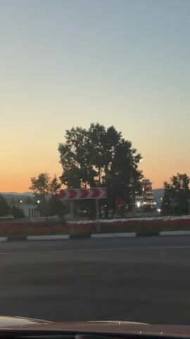 A time-lapse video shows a car driving along a road at sunset. The sky is a gradient of orange and blue. In the background, a multi-story building with a pagoda-style roof is illuminated with red and white lights. There are trees and streetlights along the road. Several cars are visible on the road, and some appear to be driving in the opposite direction. The video appears to be shot from inside another vehicle.