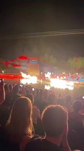 At night, a crowd of people watch a show performed on a stage in a pond. The crowd is taking pictures with their phones. The stage is lit up, and there are performers dressed in red, yellow and black uniforms. They are wearing hats and carrying swords and sticks. Flames are bursting from the stage. There are some trees in the background, and the stage is reflecting in the water. A traditional building is in the background as well.