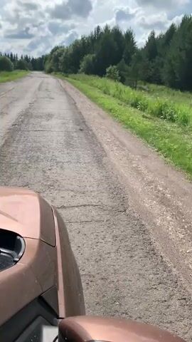 A first-person perspective video shows a car driving down a bumpy, cracked asphalt road. The road is lined with green grass and trees on either side. The sky is partly cloudy. The car's hood is visible in the lower left corner of the frame. The road appears to be in a rural or forested area.