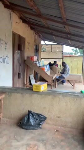 A wide shot shows an outdoor scene with a small storefront. The building has a corrugated metal roof and walls made of various materials, including some with graffiti. In the foreground, there's a wooden stool and a black trash bag on the ground. To the right, a wooden plank leans against the building.

The main action takes place at the storefront, which is partially covered by a metal gate. Inside the store, shelves are stocked with various items, likely for sale. Two young women are the central figures. The woman on the right, wearing a white fluffy jacket and a grey skirt, is facing the camera and smiling, gesturing with her hands. The woman on the left, wearing a yellow top and a patterned skirt, is partially visible inside the store and looking towards the woman in the white jacket. Another person is visible further back in the store, seated and looking towards the camera. A large brown bowl filled with what appears to be dried beans or seeds sits on a table in front of the storefront. The scene appears to be in a developing country, judging by the architecture and the goods displayed in the store.