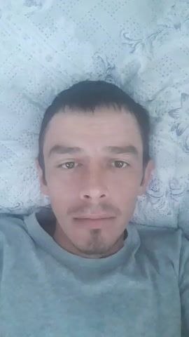 A man lies on a bed with a patterned cover and looks directly into the camera. He is wearing a gray sweatshirt. The video captures his facial expressions as he speaks.