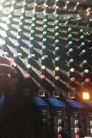 The video shows a close-up, top-down view of a sound mixing board. A person's hand is visible, manipulating several knobs on the board. The lighting is dim, with a strong light source coming from the upper left, casting shadows across the mixer. Some of the mixer's controls, like the faders and buttons, are visible in the lower part of the frame, along with some illuminated indicators labeled "PEAK". The hand appears to be adjusting multiple channels on the mixing board. The focus is on the hand and the mixer controls.