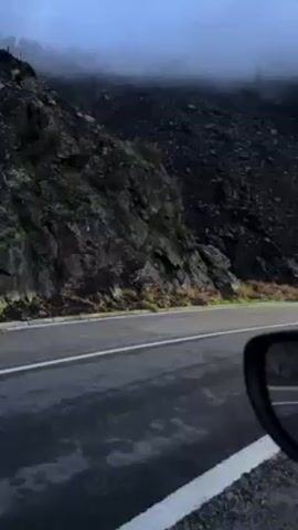 The video shows cars passing by on a mountain road. The road is wet and has white lane markings. A rocky hillside rises on the left side of the road, partially obscured by fog. A car's side mirror is visible in the lower right of the frame. Two cars pass by in the opposite direction. The sky is overcast and gray.