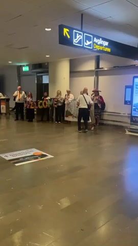 The video captures a welcoming scene at an airport, likely Stockholm-Arlanda Airport in Sweden, given the "Avgångar Departures" sign. A group of people, some dressed in traditional Swedish folk costumes, are holding up large letters spelling "WELCOME." They appear to be greeting travelers. One man in a white shirt and orange tie is dancing with a woman in a folk costume, suggesting a joyful and festive atmosphere. Another banner behind them reads "THANK YOU FOR VISITING US." Other individuals are visible in the background, some with luggage, indicating the setting is a public area of the airport. The lighting suggests it could be daytime. The overall mood conveyed is one of warmth and hospitality.