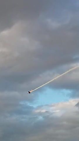 A single-engine airplane performs an aerial maneuver, leaving a white contrail against a cloudy sky. The airplane is seen from below and to the side, with the contrail creating a distinct curved line. The lighting suggests it might be late afternoon or early morning, with the sun casting a warm glow on the airplane's fuselage. The clouds are a mix of gray and white, with some patches of blue sky visible. The airplane appears to be in a banking turn, as indicated by the angled contrail.